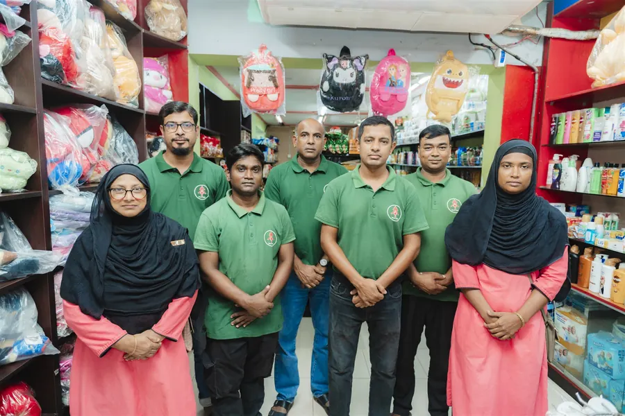 Nine members of the Nanking Bazar team in green shirts and pink uniforms, standing among the toy and cosmetics shelves