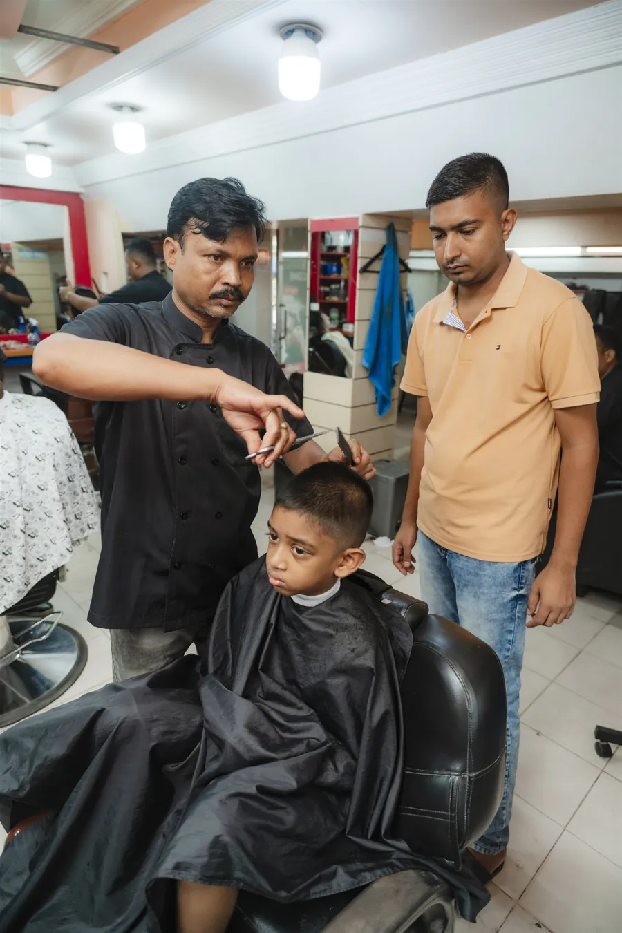 A barber cutting a boy’s hair while his father watches