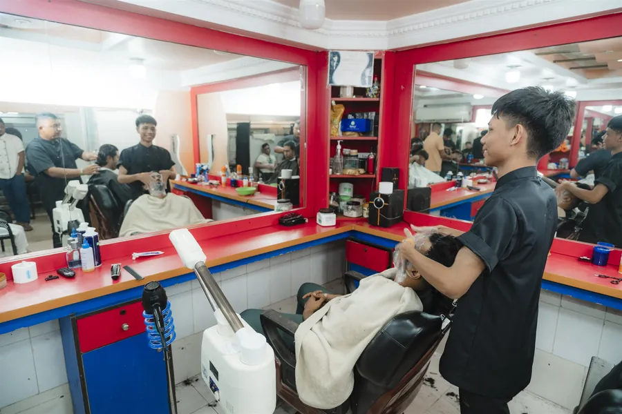 A customer reclining for face care beside a steamer while a barber works