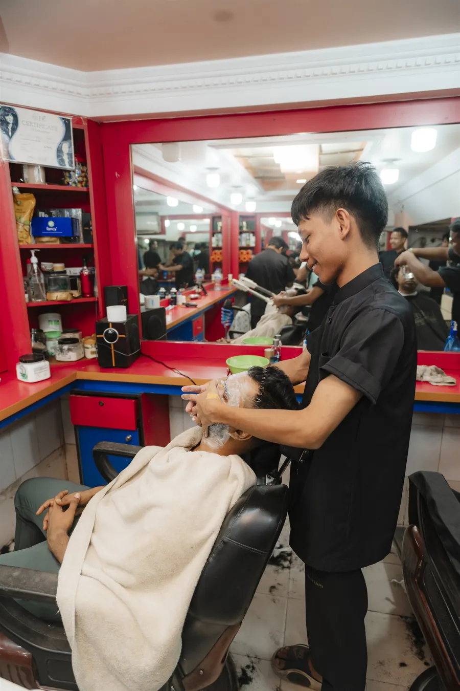 A barber working on a reclined customer’s face, the customer under a towel